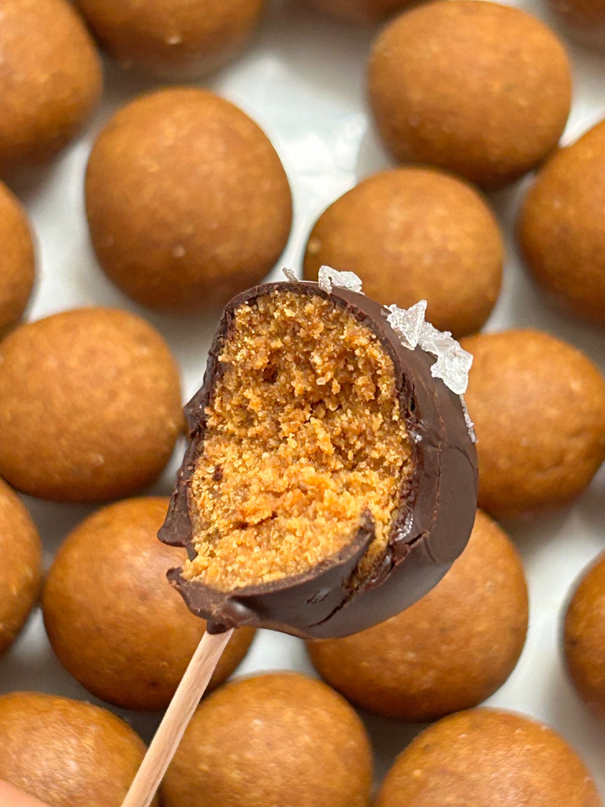 A chocolate-dipped laddoo with a bite revealing its golden center, over a tray of laddoos