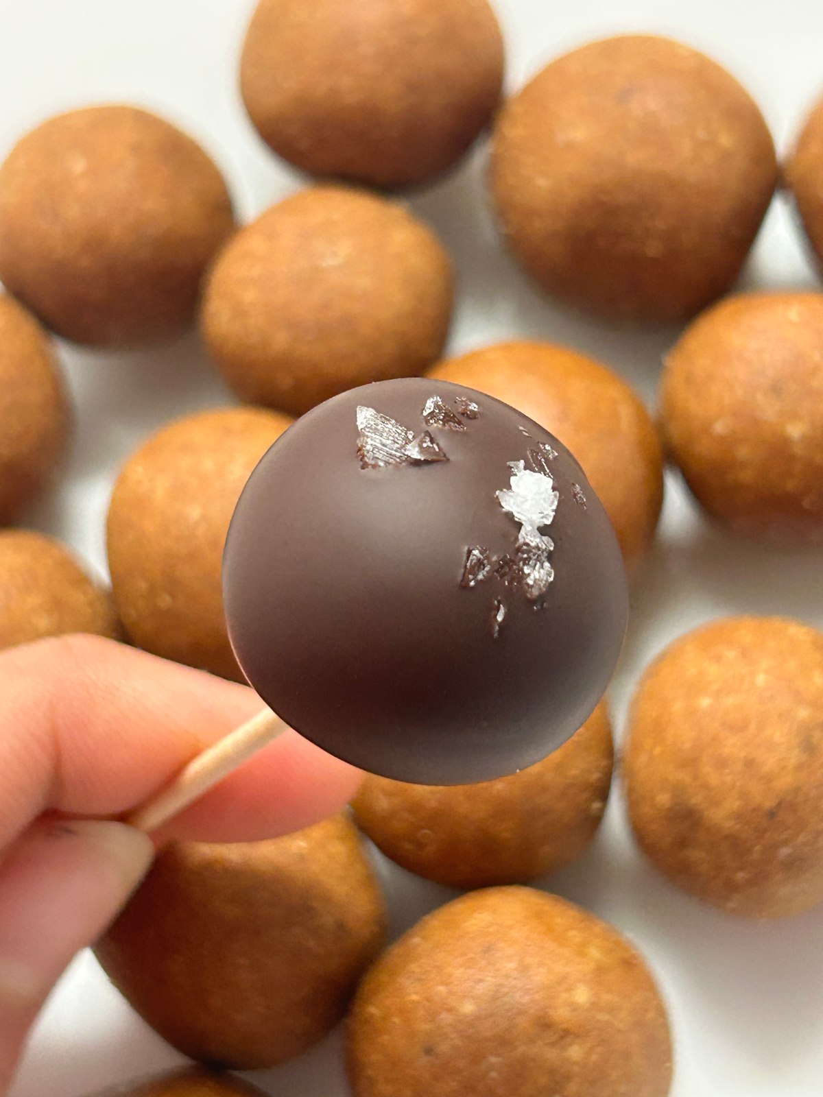 A chocolate-coated laddoo with silver garnish, held above a tray of golden laddoos